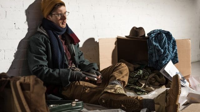homeless man sitting by brick wall surrounded by rubbish