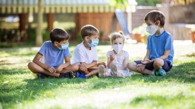Young children in protective masks on faces outdoors. Quarantine. Kids wearing safety masks while sitting on grass in park. Coronavirus prevention.