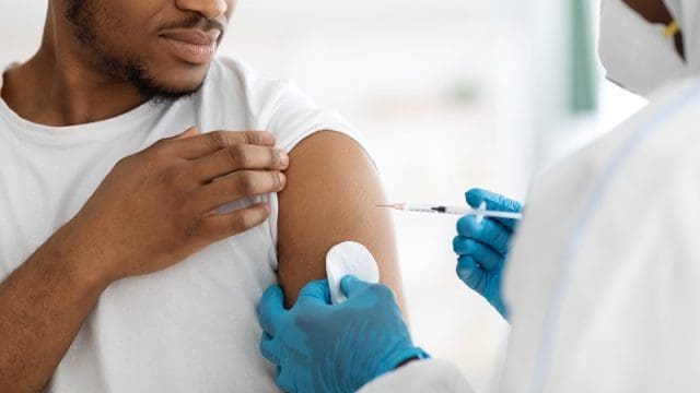 Vaccination and immunization concept. Cropped of young black man receiving vaccine shot during coronavirus pandemic. Closeup of doctor in PPE making injection in shoulder for male patient