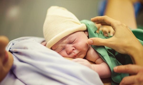 Cute newborn baby in hat sleeping on crop anonymous mother lying in delivery room after giving birth in maternity hospital
