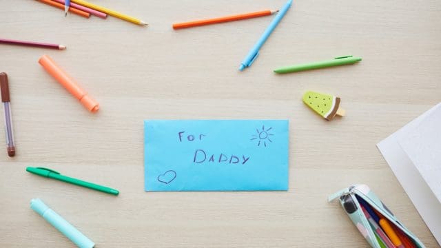 Top view background image of letter for dad with handmade gift on Fathers day set on table with colored crayons, copy space