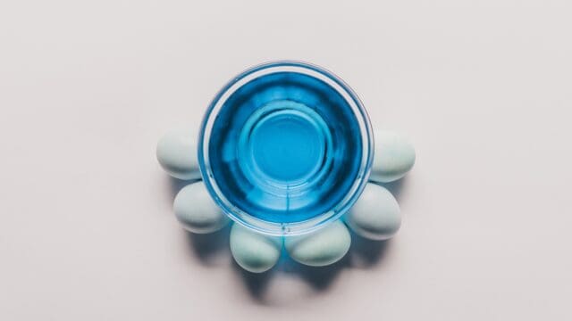 top view of glass with blue paint and chicken eggs on white surface, easter concept